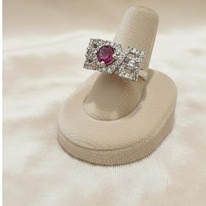 Peut inclure: Une bague en argent avec une pierre centrale ronde rose foncé, entourée de petites pierres claires. La bague présente un design géométrique avec des formes rectangulaires et carrées, présentée sur un présentoir beige.