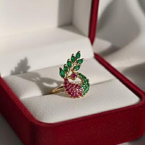 Könnte beinhalten: Ein goldfarbener Ring mit Blatt- und Wirbeldesign, besetzt mit grünen und roten Edelsteinen. Der Ring wird in einer roten Schmuckschatulle mit weißem Innenraum präsentiert. Die Edelsteine sind smaragdgrün und rubinrot.