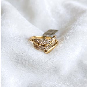 Könnte beinhalten: Ein goldfarbener Ring mit einem blattartigen Design. Der Ring hat ein Band, das sich um den Finger wickelt, und ein blattförmiges Element, das mit kleinen, klaren Edelsteinen besetzt ist. Der Ring ist vor einem weißen, strukturierten Hintergrund platziert.