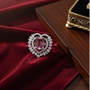 Könnte beinhalten: Ein silberner Ring mit herzförmigem Design. Der Ring hat einen großen, rechteckigen rosa Edelstein in der Mitte, umgeben von kleineren, klaren Edelsteinen. Der Ring ist vor einem tiefroten Samthintergrund platziert.