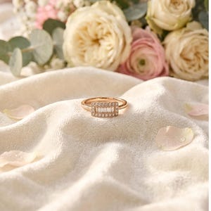 Könnte beinhalten: Ein roségoldener Ring mit einem rechteckigen Cluster aus kleinen, klaren Edelsteinen. Der Ring liegt auf einem weichen, weißen Stoff mit verstreuten Blütenblättern und einem verschwommenen Blumenarrangement im Hintergrund.