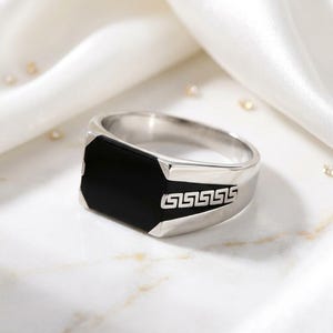 Könnte beinhalten: Ein silberfarbener Ring mit einer rechteckigen schwarzen Einlage auf der Vorderseite. Die Seiten des Rings sind mit einem silbernen griechischen Mäandermuster auf schwarzem Grund verziert, das einen Kontrast zur polierten Oberfläche bildet.