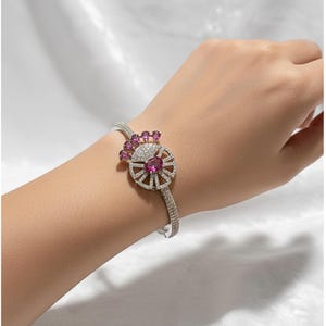 Peut inclure: Un bracelet argenté avec un motif floral orné d'une grosse pierre rose ovale au centre, entourée de petites pierres roses et de cristaux transparents. Le bracelet est posé sur un fond blanc.