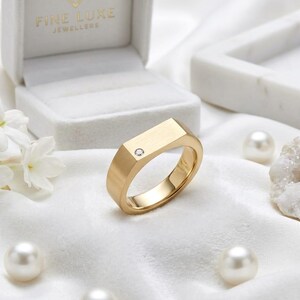 Könnte beinhalten: Ein goldener Siegelring mit matter Oberfläche und einem einzelnen kleinen runden Diamanten, der in die flache Oberseite eingelassen ist. Der Ring liegt neben einer weißen Schmuckschatulle mit der Aufschrift Fine Luxe Jewellers, weißen Blüten, Perlen und einer weißen Geode.