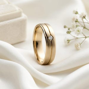 Könnte beinhalten: Ein goldener Ring mit gebürstetem, mattem Finish und einem einzelnen, kleinen, quadratisch gefassten, klaren Edelstein, der in einer vertikalen Rille auf der Außenseite des Metallbandes sitzt und für ein klares, strukturiertes Design sorgt.