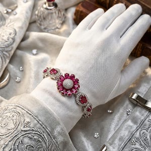 Könnte beinhalten: Ein silbernes Armband mit floralem Design. Das Armband hat eine große zentrale Blume mit rosa Blütenblättern und einem weißen Zentrum. Zusätzliche rosa Edelsteine und kleine, klare Steine sind in das Armband eingesetzt. Das Armband wird auf einem weißen Handschuh präsentiert.