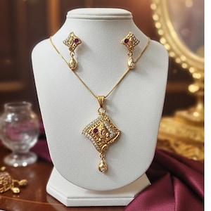 Könnte beinhalten: Ein goldfarbenes Schmuckset mit einer Anhängerkette und passenden Ohrringen. Der Anhänger und die Ohrringe haben ein rautenförmiges Design mit einem zentralen roten Stein und umliegenden klaren Kristallen. Das Set wird auf einer weißen Schmuckbüste präsentiert.