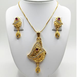 Könnte beinhalten: Ein goldfarbenes Schmuckset mit einer Anhängerkette und passenden Ohrringen. Der Anhänger und die Ohrringe haben ein florales Design mit roten Edelsteinen und klaren Kristallen. Die Halskette hat eine feine Kette.