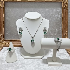 May include: A jewelry set featuring a necklace, earrings, ring, and bracelet. The pieces have a silver finish and are adorned with emerald-green teardrop-shaped stones and clear accent stones. The jewelry is displayed on white mannequins and stands.