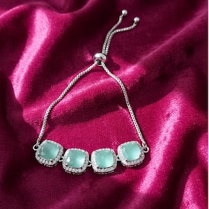 Peut inclure: Bracelet argenté orné de cinq pierres carrées vert clair. Chaque pierre est entourée d'un halo de petits cristaux transparents. Le bracelet est doté d'une chaîne réglable et est présenté sur un fond de velours rose foncé.