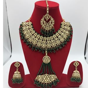 Könnte beinhalten: Ein aufwendiges, goldfarbenes Schmuckset mit grünen Akzenten, präsentiert auf einer roten Schaufensterpuppe. Das Set umfasst eine Halskette, Ohrringe und ein Kopfstück. Die Halskette hat ein großes, geschwungenes Design mit komplizierten Details und grünen Perlen.