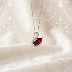 May include: A silver-toned necklace featuring a swan-shaped pendant. The swan's body is a large, pear-shaped deep red gemstone, while its neck and head are outlined with small, clear, sparkling stones set into the metallic frame.