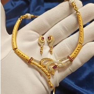 Könnte beinhalten: Ein goldfarbenes Halsketten- und Ohrring-Set. Die Halskette hat ein geschwungenes Design mit eingebetteten klaren und roten Edelsteinen. Die Ohrringe haben ein ähnliches Design mit einem hängenden roten Edelstein. Der Schmuck wird auf einer weißen Oberfläche präsentiert.