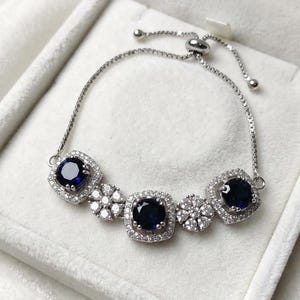 Könnte beinhalten: Silberarmband mit fünf dunkelblauen, quadratisch geschliffenen Edelsteinen. Das Armband hat eine filigrane Kette und befindet sich in einer weißen Samt-Schmuckschatulle. Die Edelsteine sind von kleineren, klaren Steinen umgeben.
