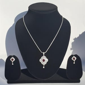 Könnte beinhalten: Ein silbernes Halsketten- und Ohrring-Set, präsentiert auf einem schwarzen Schmuckständer. Die Halskette hat einen Anhänger mit einem roten Edelstein in einem vierblättrigen Kleeblatt-Design. Die Ohrringe haben das gleiche Design.