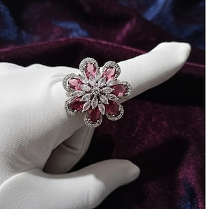 Könnte beinhalten: Silberner Ring in Blumenform mit rosa und klaren Edelsteinen. Die Blütenblätter sind tiefrosa und die Mitte ist ein Cluster aus klaren Steinen. Der Ring ist vor einem weißen Hintergrund platziert.