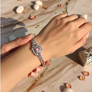 Puede incluir: Una pulsera plateada con una piedra rosa central en forma de pera, rodeada de piedras claras más pequeñas. La pulsera tiene un diseño abierto con detalles intrincados. La imagen muestra la pulsera en una muñeca, con una superficie similar al mármol.