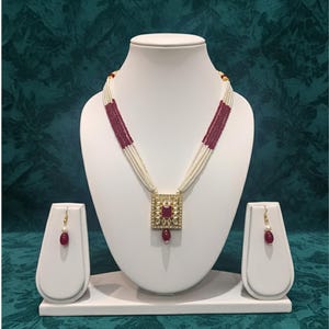 Könnte beinhalten: Ein goldfarbenes Halsketten- und Ohrring-Set, das auf einer weißen Büste ausgestellt ist. Die Halskette hat mehrere Stränge aus weißen Perlen, die mit Abschnitten aus weinroten Perlen durchsetzt sind. Der Anhänger und die Ohrringe haben einen quadratischen weinroten Stein.