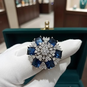 May include: A silver ring with a floral design. It features six large, rectangular, deep blue gemstones and a centre of smaller, clear stones. The ring is held against a white glove. The ring's design is reminiscent of a flower.