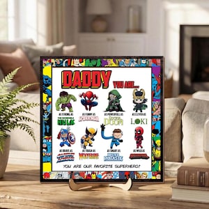 Può includere: Stampa incorniciata a tema supereroi. La stampa presenta la parola "DADDY" a lettere grandi, insieme a vari personaggi Marvel e frasi come "Forte come Hulk" e "Sei il nostro supereroe preferito!" La cornice è nera.