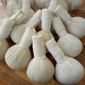 May include: A collection of beige, round, herbal massage compress balls. Each ball has a cylindrical handle wrapped with white twine. The balls are made of a textured fabric and are arranged in a pile.