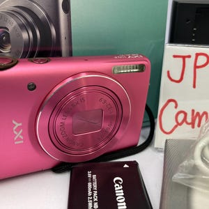 May include: A pink Canon IXY digital camera with a zoom lens. The camera has a black wrist strap and a battery pack. The image also shows a box with the text "JP Cam" written on it.