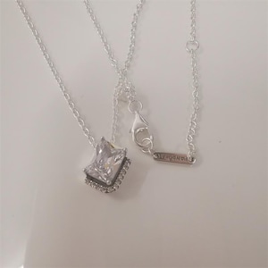 Peut inclure: Un collier argenté avec un pendentif carré en pierre précieuse transparente. Le pendentif est entouré de petites pierres transparentes. Le collier a une chaîne délicate et un fermoir. La marque "PANDORA" est visible sur une petite étiquette.