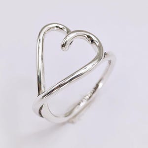 May include: A silver ring in the shape of a heart. The ring is made of a shiny, polished metal and features a unique open design. The heart shape is formed by two curved lines that meet at the top and cross at the bottom.