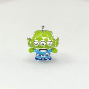 May include: A small, three-eyed green alien figurine with a silver antenna. The alien is wearing a blue uniform with a silver buckle. The figurine is a collectible item, likely from a popular animated film.