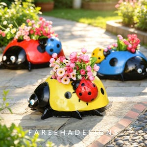 May include: Three ladybug-shaped planters in red, yellow, and blue, each featuring black spots and a black head with white eyes. The planters are filled with small pink and white artificial flowers. The text FAREEHADESIGNS is visible at the bottom.