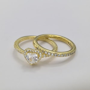 May include: A gold-colored ring set. The engagement ring features a heart-shaped, clear gemstone surrounded by smaller stones. The wedding band is a simple gold band with small stones. Both rings are on a white surface.