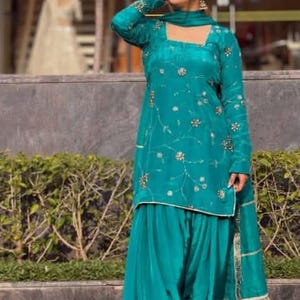 May include: A teal long-sleeved tunic with a square neckline and gold floral embroidery paired with matching pleated salwar trousers and a coordinating dupatta. The ensemble features a smooth, satin-like finish and gold trim on the dupatta edges.