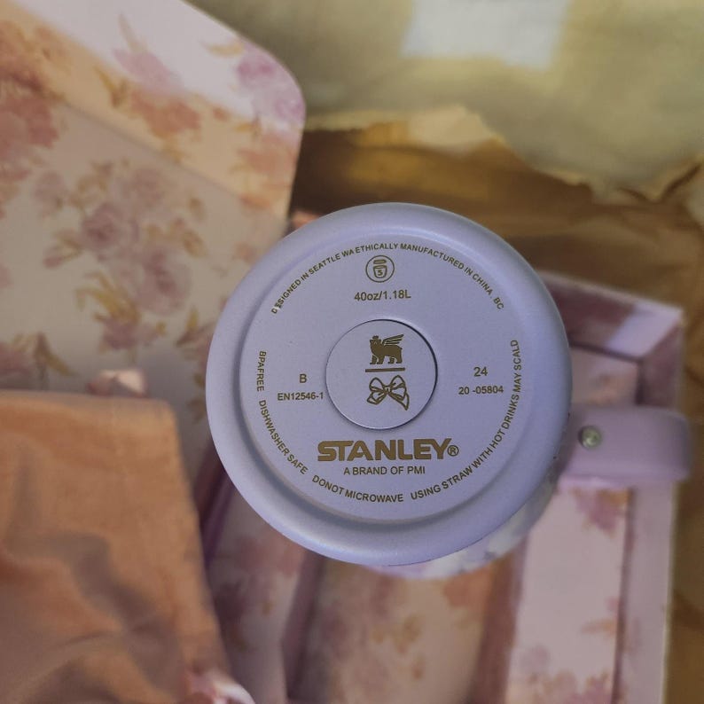 K&ouml;nnte beinhalten: Der Boden eines hellvioletten Stanley-Bechers mit der Aufschrift 40oz/1.18L, einem Logo mit einem gefl&uuml;gelten Wesen und einer Schleife sowie dem Markennamen Stanley. Der Text enth&auml;lt Hinweise wie sp&uuml;lmaschinenfest und nicht mikrowellengeeignet.