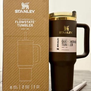 May include: A dark brown Stanley Quencher H2.0 FlowState tumbler with a 40 oz capacity, featuring a matching handle, a clear lid, and a straw. The tumbler is displayed next to its cardboard packaging, which lists 11 hours cold, 2 days iced, and 7 hours hot.
