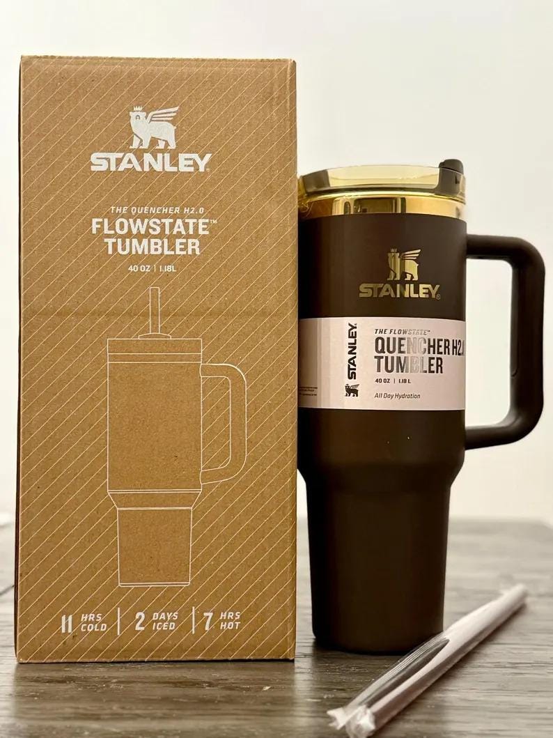 May include: A dark brown Stanley Quencher H2.0 FlowState tumbler with a 40 oz capacity, featuring a matching handle, a clear lid, and a straw. The tumbler is displayed next to its cardboard packaging, which lists 11 hours cold, 2 days iced, and 7 hours hot.