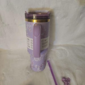 K&ouml;nnte beinhalten: Ein hellvioletter Isolierbecher mit Blumenmuster, goldenem Rand und passendem violettem Henkel. Der Becher verf&uuml;gt &uuml;ber einen transparenten Deckel, einen vertikalen Griff und ein Produktinformationsetikett an der Seite. Ein violetter Strohhalm liegt bei.