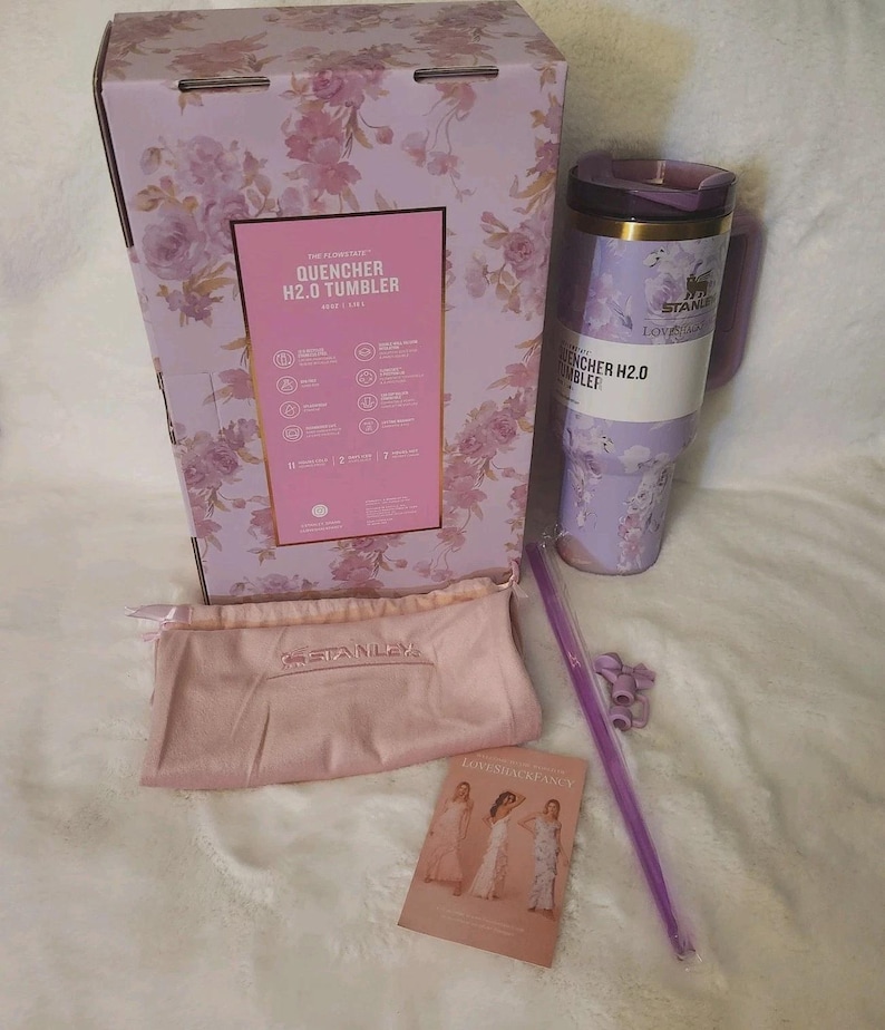 K&ouml;nnte beinhalten: Ein Stanley Quencher H2.0 Becher mit hellviolettem Blumenmuster, dazu die passende Schachtel, ein hellrosa Kordelzugbeutel mit Stanley-Logo, ein langer violetter Strohhalm, zwei kleine violette Strohhalmaufs&auml;tze und eine Infokarte.
