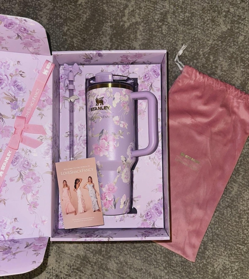 K&ouml;nnte beinhalten: Ein lavendelfarbener Stanley-Becher mit Blumenmuster, ein passender Strohhalm mit Schleifenaufsatz und eine kleine Karte mit dem Text LoveShackFancy, alles in einer blumigen Geschenkbox neben einem rosa Samtbeutel mit Kordelzug.