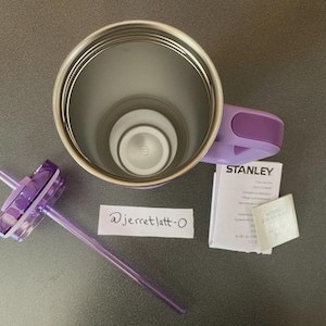 May include: A lavender Stanley tumbler with a metallic interior, a matching purple handle, and a clear lid with a purple straw. Included are a small white instruction booklet with the Stanley logo and a handwritten note reading @jerretlatt-0.
