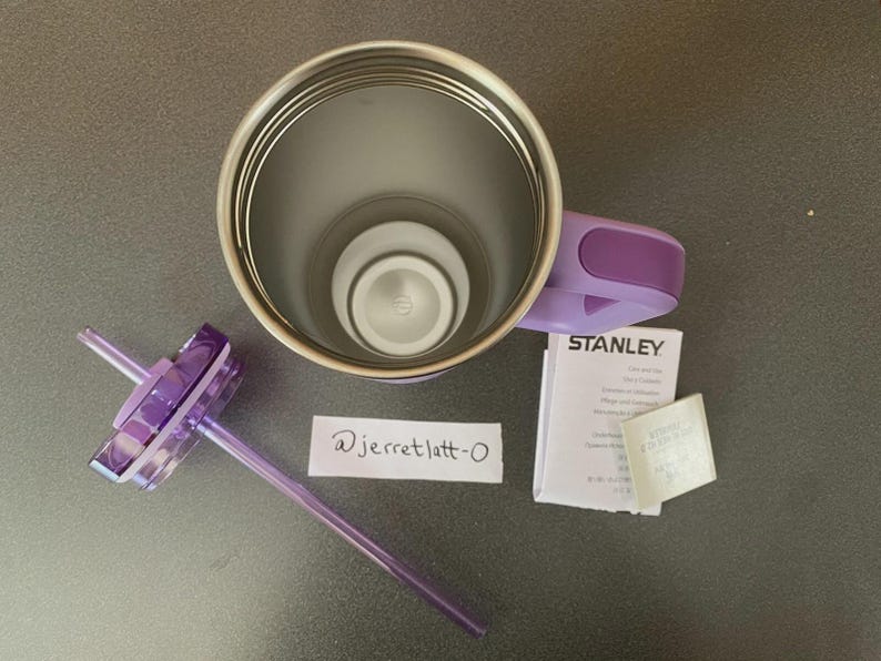 May include: A lavender Stanley tumbler with a metallic interior, a matching purple handle, and a clear lid with a purple straw. Included are a small white instruction booklet with the Stanley logo and a handwritten note reading @jerretlatt-0.