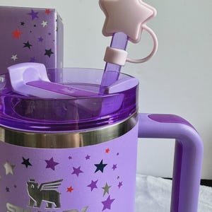 May include: A light purple Stanley tumbler featuring a silver bear logo and a pattern of multi-colored stars. The tumbler has a matching purple handle, a translucent purple lid, and a straw topper shaped like a pale pink five-pointed star.