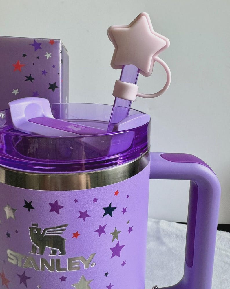 May include: A light purple Stanley tumbler featuring a silver bear logo and a pattern of multi-colored stars. The tumbler has a matching purple handle, a translucent purple lid, and a straw topper shaped like a pale pink five-pointed star.
