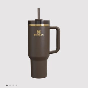 May include: A dark brown insulated tumbler with a matching handle and a dark brown lid featuring a central straw. The cup displays a gold Stanley logo with a winged bear emblem on the upper front section, accented by a thin gold band below the lid.