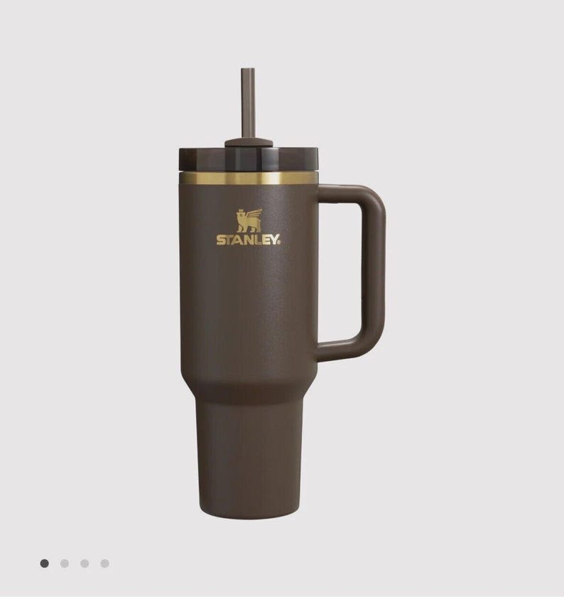 May include: A dark brown insulated tumbler with a matching handle and a dark brown lid featuring a central straw. The cup displays a gold Stanley logo with a winged bear emblem on the upper front section, accented by a thin gold band below the lid.
