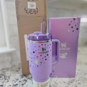 May include: A light purple Stanley Quencher tumbler with a handle, a clear straw, and a lid. The exterior features a pattern of small black, red, and silver stars. The Stanley logo is printed on the front, and a product label is attached to the base.