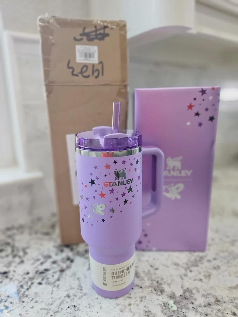 May include: A light purple Stanley Quencher tumbler with a handle, a clear straw, and a lid. The exterior features a pattern of small black, red, and silver stars. The Stanley logo is printed on the front, and a product label is attached to the base.