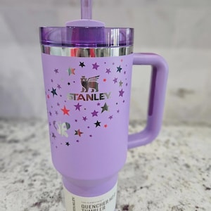 May include: A light purple Stanley Quencher H2.0 FlowState tumbler with a handle, a purple lid, and a matching straw. The exterior features a metallic silver bear logo, the text STANLEY, and a pattern of small stars and a butterfly in various colors.
