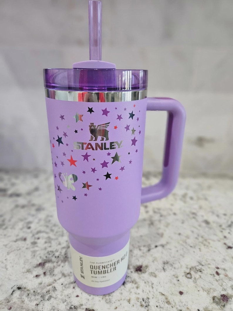 May include: A light purple Stanley Quencher H2.0 FlowState tumbler with a handle, a purple lid, and a matching straw. The exterior features a metallic silver bear logo, the text STANLEY, and a pattern of small stars and a butterfly in various colors.