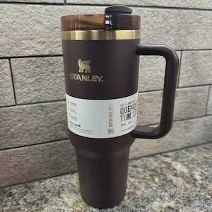 May include: A dark brown Stanley FlowState Quencher tumbler with a 40 oz capacity, featuring a gold-tone rim, a matching dark brown handle, and a translucent dark brown lid. A white product label is wrapped around the center of the tumbler.