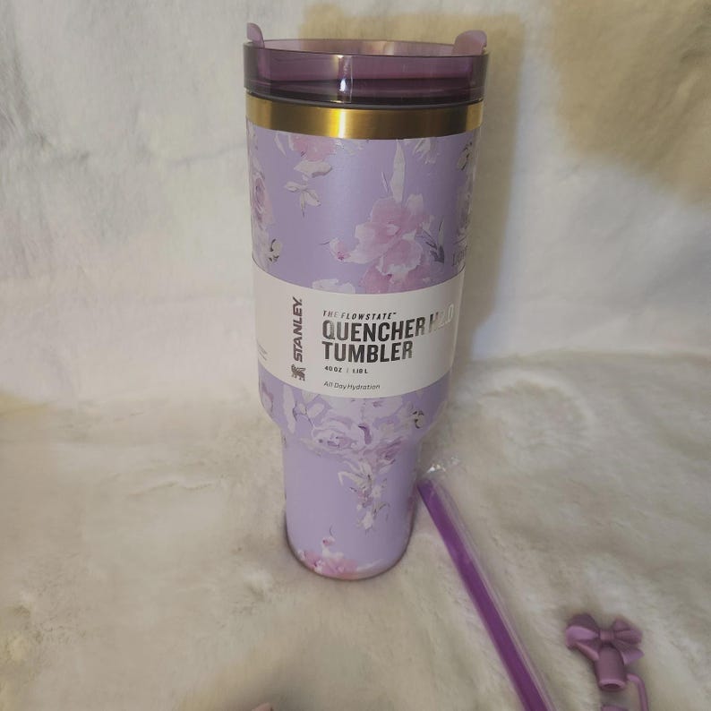 K&ouml;nnte beinhalten: Ein hellvioletter Stanley Flowstate Quencher H2.0 Becher mit Blumenmuster, goldenem Rand und durchsichtigem violettem Deckel. Das Etikett zeigt The Flowstate Quencher H2.0 Tumbler 40 oz | 1,18 L. Enth&auml;lt einen violetten Strohhalm und Schleifenanh&auml;nger.